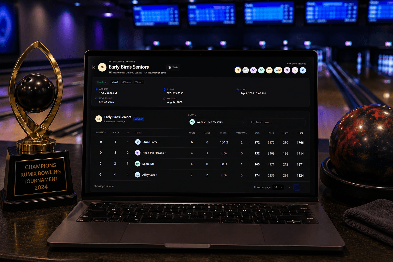 Laptop on a bowling centre counter showing a league's public standings page, next to a tournament trophy and a bowling ball